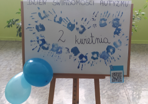 Na zdjęciu widoczny jest plakat przygotowany przez uczniów klasy 2a. Na plakacie znajdują się odciśnięte dłonie w kolorze niebieskim. Kompozycja przedstawia niebieskie łapki w kształcie serca.